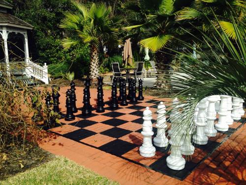 A large outdoor chess set with black and white pieces on a checkered board, surrounded by palm trees and a gazebo.