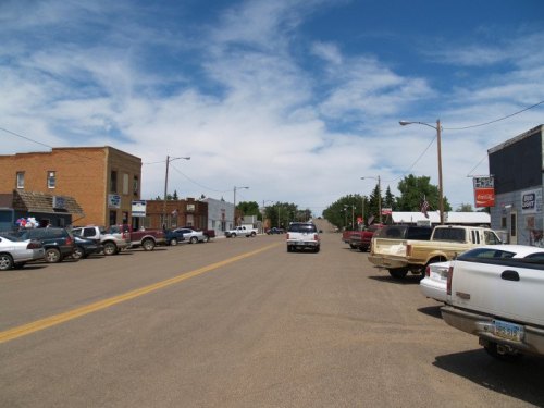 A small town street lined with parked cars, shops, and a clear blue sky with scattered clouds.