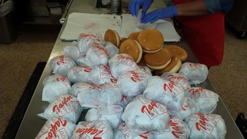 A countertop with wrapped sandwiches and burger buns, with a person in blue gloves preparing food in the background.