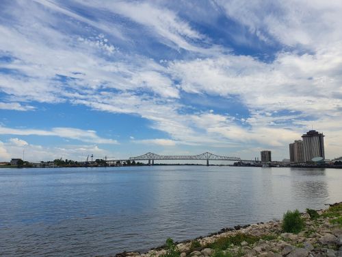 A river scene featuring a bridge, city skyline, and blue sky with clouds, viewed from a rocky shoreline.
