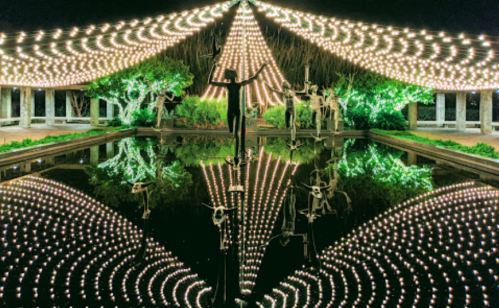 A serene night scene with illuminated trees and sculptures reflected in a calm pond, creating a magical atmosphere.