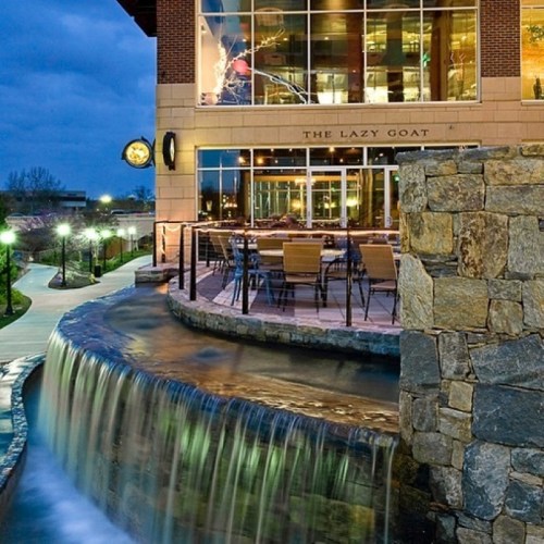 A modern restaurant with outdoor seating beside a flowing water feature, set against a twilight sky.