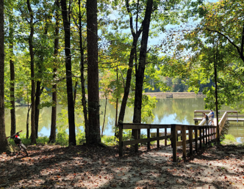 A serene lake view surrounded by trees, with a wooden path leading to a dock and two people fishing.