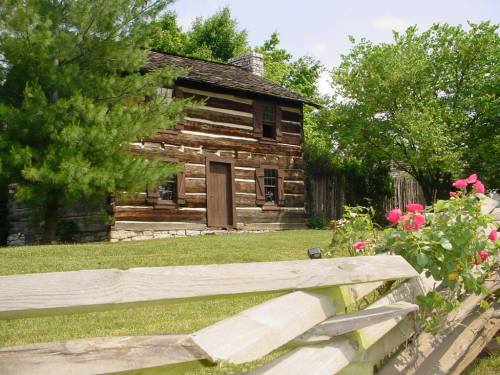 A rustic log cabin surrounded by greenery and flowers, with a wooden fence in the foreground.