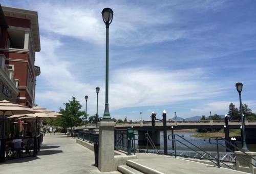 A riverside walkway with a bridge in the background, lined with trees and street lamps under a blue sky.
