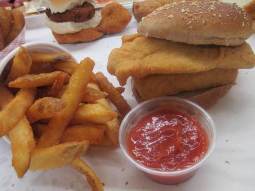 A plate featuring fried fish fillets stacked on a bun, served with French fries and a small cup of ketchup.