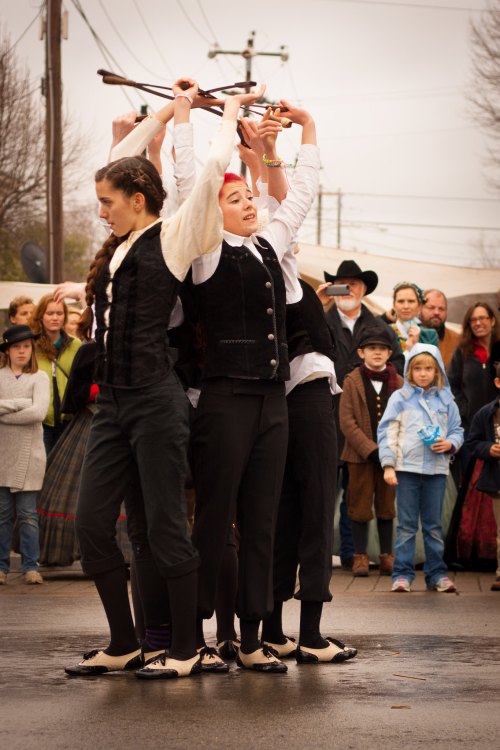 A group of performers in historical costumes are dancing with props, while an audience watches in the background.