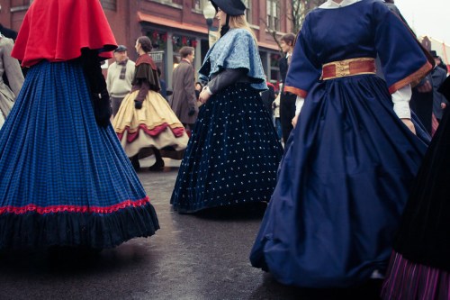 People in historical dresses with colorful skirts walking in a busy street during a cultural event.