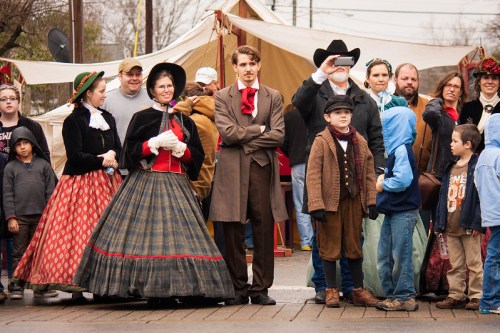 A group of people in historical costumes stands outdoors, with tents in the background and a mix of adults and children.