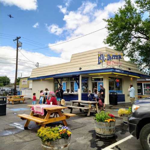 A busy ice cream shop with families enjoying treats outside, picnic tables, and a clear blue sky overhead.