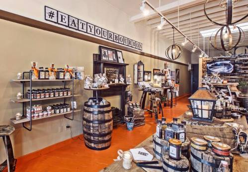 A cozy shop interior featuring wooden barrels, shelves of products, and a "#EATYOURBOURBONS" sign on the wall.
