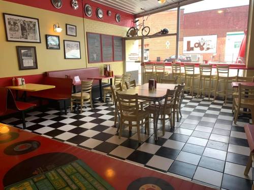 A retro diner interior with checkered floors, red booths, and vintage decor, featuring tables and large windows.