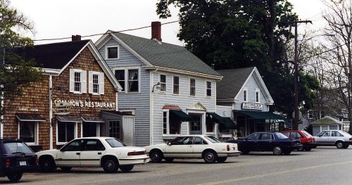 A street view of a small town featuring several restaurants and parked cars on a cloudy day.