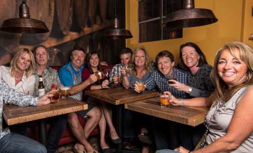 A group of ten people smiling and holding drinks at a cozy bar with warm lighting and wooden tables.