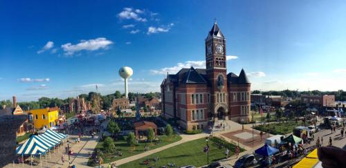 A vibrant town square featuring a historic clock tower, colorful tents, and people enjoying a sunny day.
