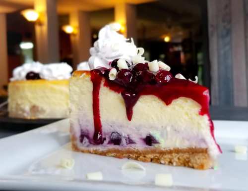 A slice of cheesecake topped with berry sauce, whipped cream, and white chocolate shavings on a white plate.