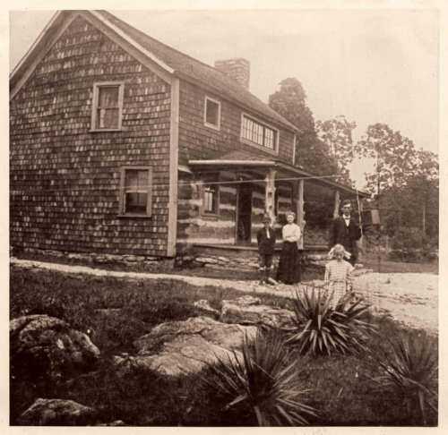 A vintage photo of a wooden house with a family standing outside, surrounded by rocks and greenery.