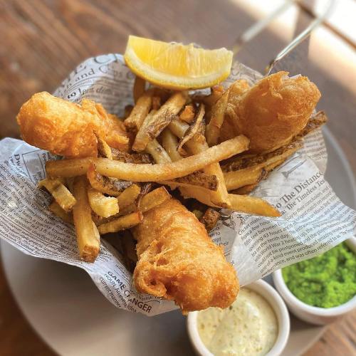 A serving of crispy fish and chips with a lemon wedge, accompanied by tartar sauce and green mushy peas.