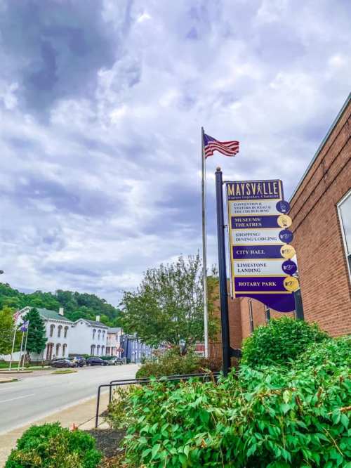 Signpost in Maysville, KY, displaying local attractions and city hall, with a cloudy sky and American flag in the background.