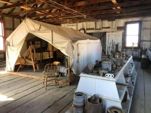 Interior of a rustic building featuring a canvas tent, wooden furniture, and various vintage tools and artifacts.