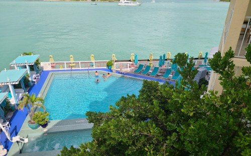 Aerial view of a pool surrounded by lounge chairs, with a turquoise water backdrop and boats in the distance.
