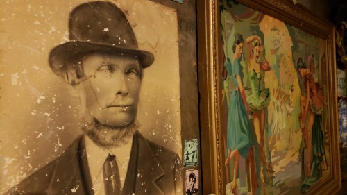 A vintage portrait of a man in a hat next to a colorful painting of women in dresses, set in a rustic environment.