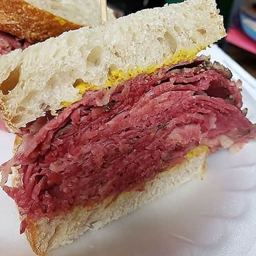 Close-up of a sandwich with layers of pink pastrami and yellow mustard on soft white bread.