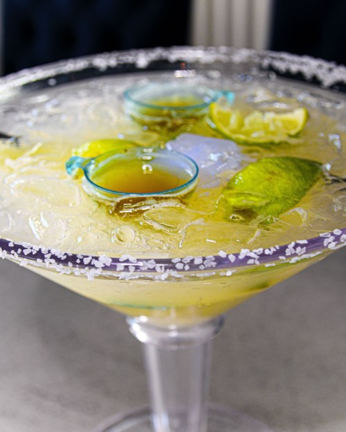 A large margarita glass filled with ice, lime slices, and two small shot glasses on top, rimmed with salt.