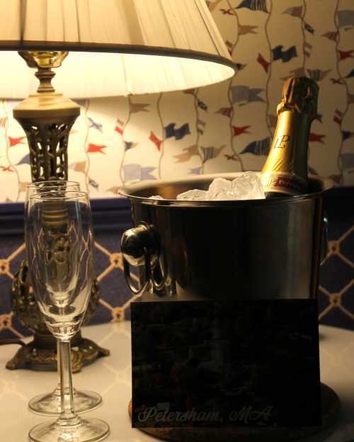 A champagne bottle in an ice bucket beside two flutes and a lamp, with a decorative sign reading "Peterham, MA."