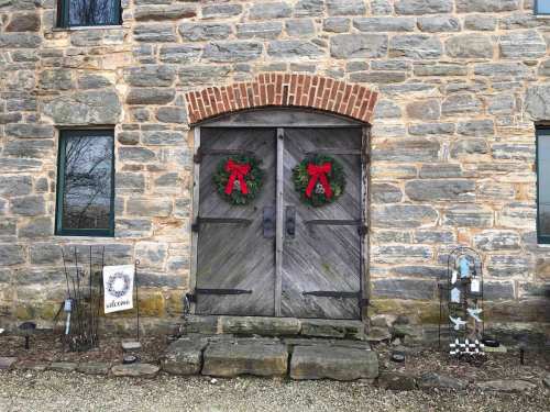 A rustic stone building with a wooden double door adorned with wreaths and a welcome sign nearby.