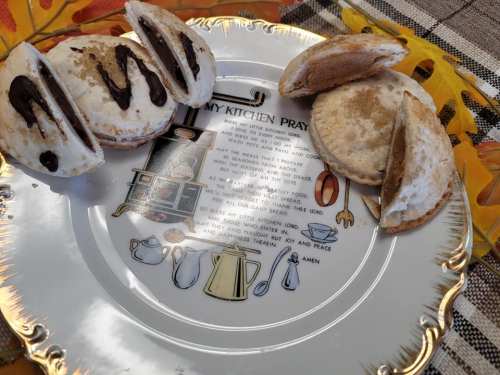 A decorative plate with a kitchen prayer and two cookies drizzled with chocolate, set on a patterned tablecloth.
