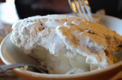 A close-up of a slice of meringue pie with a fluffy white topping and a creamy filling, served on a plate.