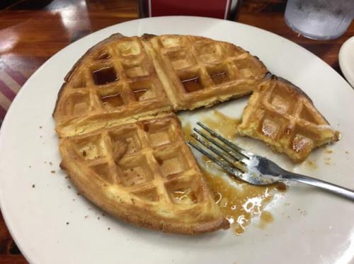 A partially eaten waffle on a plate, with syrup and a fork beside it.