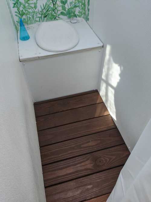 A small, simple bathroom with a wooden floor, a white toilet, and a blue bottle on a shelf. Green plants decorate the wall.