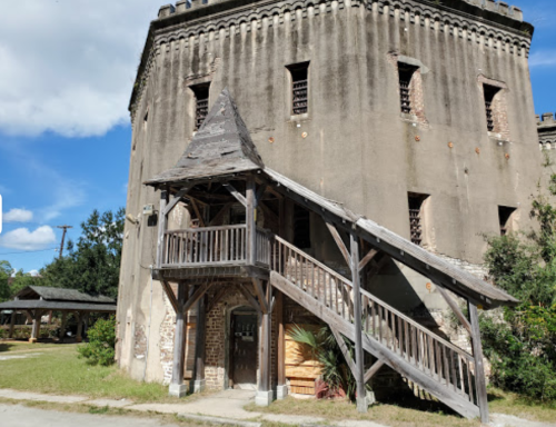 Historic building with a wooden staircase and overhang, surrounded by greenery and blue skies.