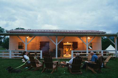 A group of people relaxes in Adirondack chairs around a fire, with a barn and cloudy sky in the background.