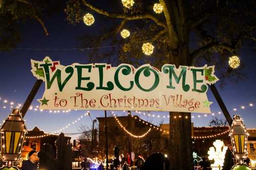 A festive sign reading "Welcome to The Christmas Village" adorned with holiday decorations and lights.