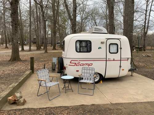 A small white camper parked on a concrete pad, surrounded by trees, with two chairs and a table set up outside.