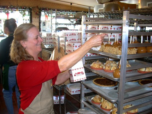 A woman in an apron smiles while placing treats on a rack in a bakery, surrounded by various baked goods.