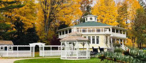 A charming yellow house with a green roof, surrounded by vibrant autumn trees and a white gazebo in the foreground.
