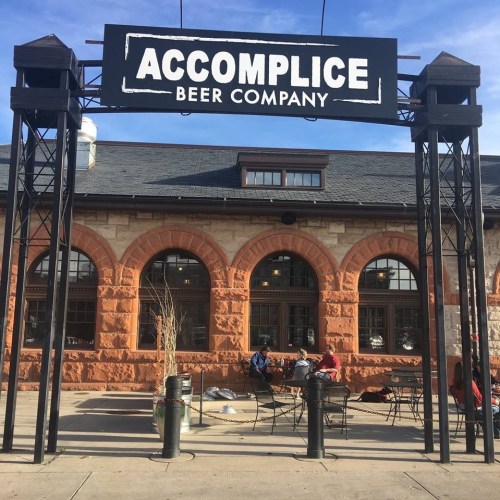 Exterior of Accomplice Beer Company, featuring a large sign and outdoor seating area with patrons enjoying the space.