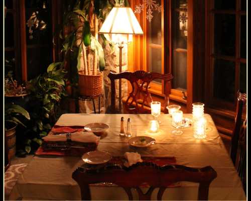 A cozy dining table set for two, illuminated by candles and a warm lamp, surrounded by plants and wooden decor.