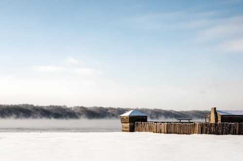 A snowy landscape with a wooden structure and a foggy river in the background under a clear blue sky.