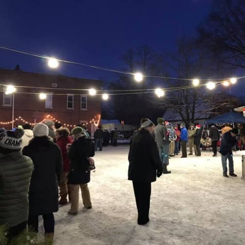 A winter evening scene with people gathered outdoors, string lights overhead, and snow on the ground.