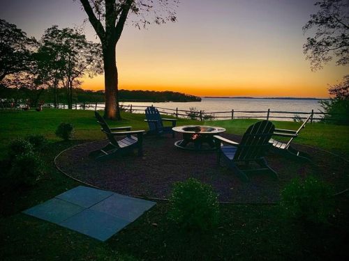A serene lakeside scene at sunset, featuring a fire pit surrounded by Adirondack chairs and lush greenery.