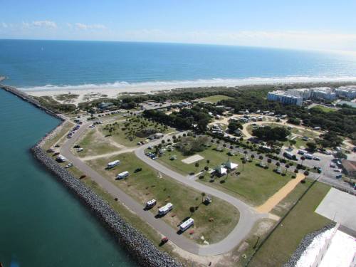 Aerial view of a coastal park with RVs, green spaces, and a beach along the ocean under a clear blue sky.