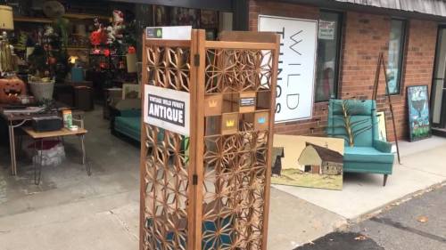 A wooden antique display stand outside a vintage shop, with various items and artwork visible in the background.