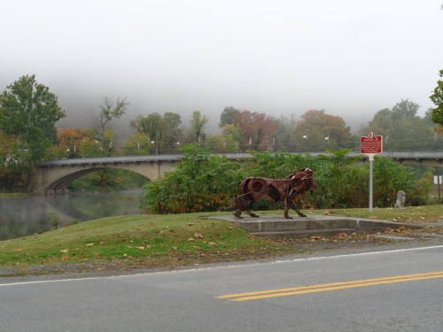 A metal sculpture of a lion stands near a riverbank, with a bridge and foggy trees in the background.
