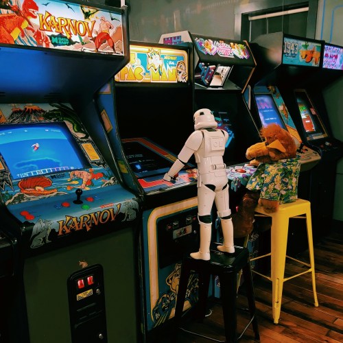 A stormtrooper figurine plays an arcade game, while a teddy bear in a dress sits nearby at another machine.
