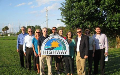 Group of people standing by a "Florida Scenic Highway" sign in a grassy area with trees and boats in the background.